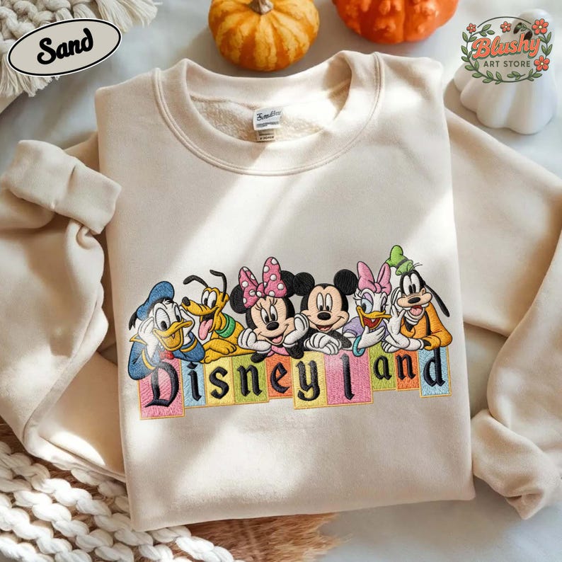 May include: A sand-colored crewneck sweatshirt featuring an embroidered graphic of Disney characters including Donald Duck, Pluto, Minnie Mouse, Mickey Mouse, Daisy Duck, and Goofy. Below the characters, the word Disneyland is stitched in black letters.