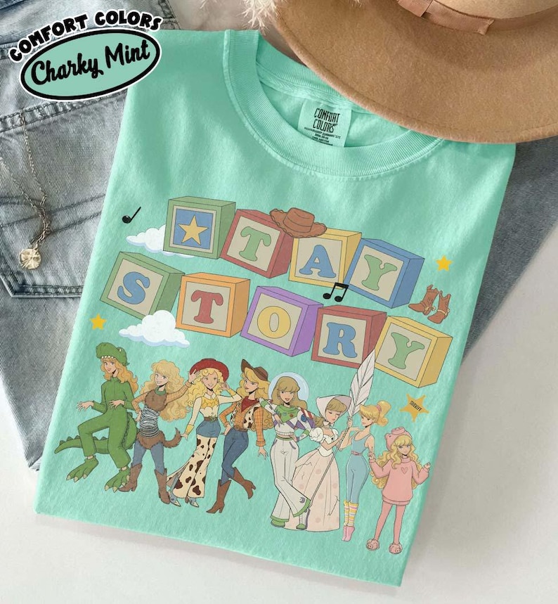 May include: A mint green t-shirt featuring a graphic of colourful alphabet blocks spelling out STAY STORY. Below the blocks are several illustrated characters dressed in various costumes, including a dinosaur, a cowgirl, and a space ranger.
