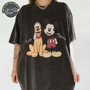 May include: Black t-shirt featuring Pluto and Mickey Mouse. Pluto is light brown with a red tongue, and Mickey Mouse is in red shorts. The shirt has the text "Pluto" and "Mickey Mouse". The shirt also has the text "Comfort Colors Pepper".