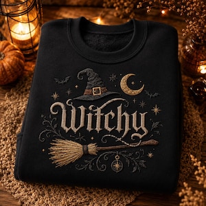 Op de afbeelding: Een zwart sweatshirt met ronde hals en een borduursel van het woord Witchy in beige letters. Het ontwerp bevat een puntige heksenhoed, een maansikkel, sterren, vleermuizen, een bezemsteel en een kompasroos in goud- en bruintinten.