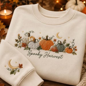 Peut inclure: Un sweat-shirt à col rond couleur crème orné de citrouilles brodées dans des tons orange, blanc et vert sauge, agrémenté de feuilles d'automne, d'étoiles et d'un croissant de lune. Le texte Spooky Harvest est brodé sous les citrouilles.