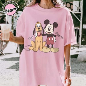 May include: Pink t-shirt featuring a graphic of Mickey Mouse and Pluto. Mickey Mouse is in black, red, and white, while Pluto is yellow. The text "Mickey Mouse" is written in cursive. The shirt has the text "Comfort Colors Blossom" in a circle.