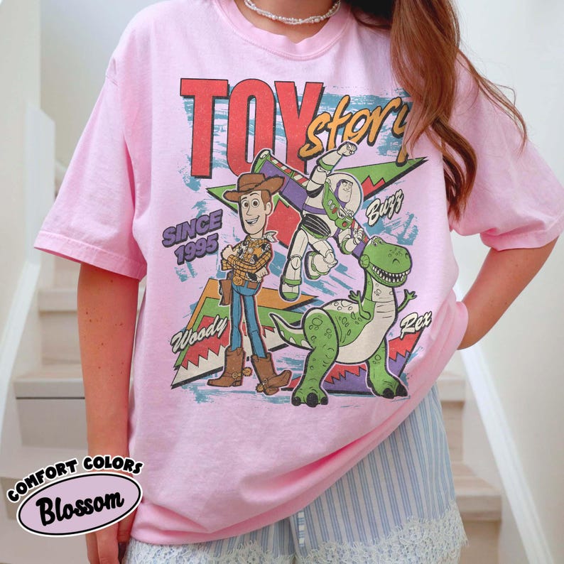 Puede incluir: Camiseta rosa claro con el logo de Toy Story y los personajes Woody, Buzz y Rex. El dise&ntilde;o incluye el texto "Since 1995". La camiseta tiene mangas cortas y cuello redondo.