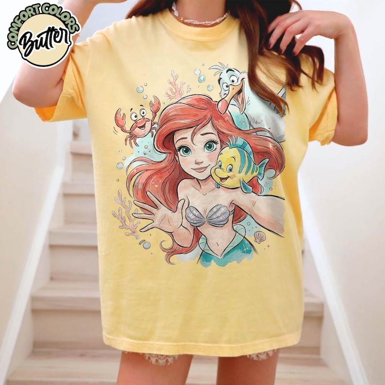 Może przedstawiać: Ż&oacute;łta koszulka z grafiką Arielki z Małej Syrenki, Florka, Sebastiana i Ptaka. Projekt jest w stylu akwareli, z logo Comfort Colors Butter w lewym g&oacute;rnym rogu.