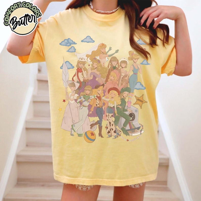 May include: A pale yellow short-sleeve t-shirt featuring a graphic print of multiple illustrated characters in various outfits, including a cowboy hat, a space suit, and a wedding dress, with blue clouds and a yellow ball, plus a Comfort Colors Butter logo.