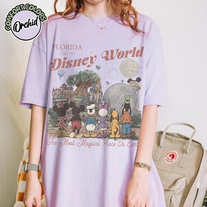 May include: A light purple t-shirt featuring a Disney World graphic. The design includes the text "FLORIDA Est. 1971 Disney World" and a colorful illustration of Disney characters and landmarks. The shirt also has the text "The Most Magical Place On Earth."