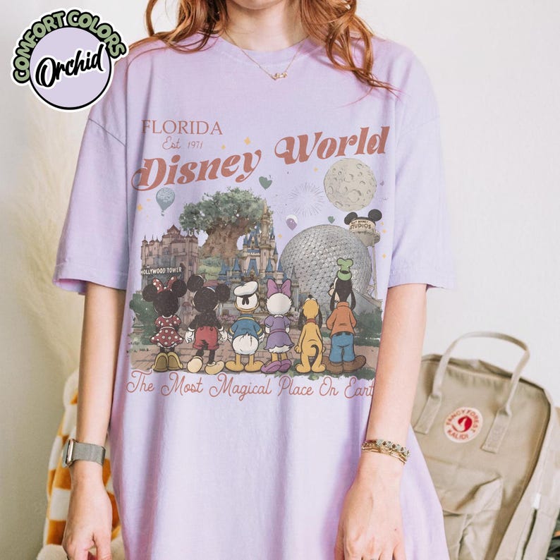 May include: A light purple t-shirt featuring a Disney World graphic. The design includes the text "FLORIDA Est. 1971 Disney World" and a colorful illustration of Disney characters and landmarks. The shirt also has the text "The Most Magical Place On Earth."