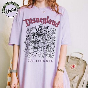 Puede incluir: Una camiseta de color lila claro con un gr&aacute;fico de Disneyland que presenta a Mickey Mouse, Minnie Mouse, Donald Duck, Daisy Duck, Goofy y Pluto. El texto "Disneyland California" est&aacute; impreso debajo de los personajes.
