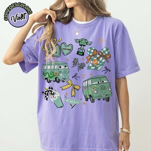 Puede incluir: Una camiseta violeta Comfort Colors con gr&aacute;ficos de dibujos animados de furgonetas verdes, corazones, lazos y un trofeo. La camiseta incluye el texto "Cars" y "Filmore". El dise&ntilde;o tiene una est&eacute;tica l&uacute;dica y caprichosa.