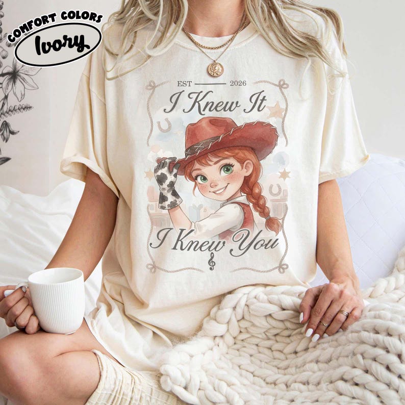 Peut inclure: Un t-shirt ivoire &agrave; manches courtes avec l'illustration d'une cow-girl de dessin anim&eacute; aux cheveux roux, portant un chapeau marron et un gant &agrave; motif vache. Le texte indique "EST 2026", "I Knew It" et "I Knew You" dans un cadre d&eacute;coratif.