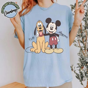 May include: Light blue t-shirt featuring Mickey Mouse and Pluto. Mickey Mouse is in black, red, and yellow, with Pluto in yellow. The shirt has the text "Mickey Mouse" and "Pluto". The shirt has the Comfort Colors Chambray logo.