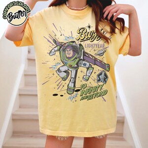 May include: Yellow t-shirt featuring a graphic of Buzz Lightyear with the text "Buzz Lightyear" and "To Infinity and Beyond." The design includes a cartoon image of Buzz Lightyear in his space suit, with a planet and starbursts.