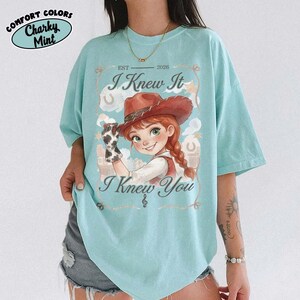 Peut inclure: Un t-shirt vert menthe clair avec une illustration de cow-girl aux cheveux roux portant un chapeau marron et un gant &agrave; motif vache. Le texte indique EST 2026, I Knew It et I Knew You, avec une petite note de musique sous le texte.