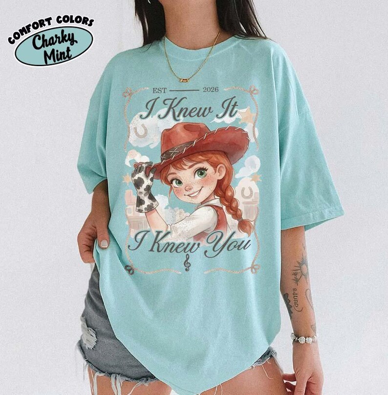 Peut inclure: Un t-shirt vert menthe clair avec une illustration de cow-girl aux cheveux roux portant un chapeau marron et un gant &agrave; motif vache. Le texte indique EST 2026, I Knew It et I Knew You, avec une petite note de musique sous le texte.