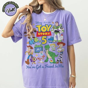 May include: A light purple short-sleeve t-shirt featuring a graphic print of Disney Pixar Toy Story characters including Woody, Buzz Lightyear, Jessie, Rex, and Forky. The text reads Toy Story 5 and You've Got a Friend In Me with a small logo.