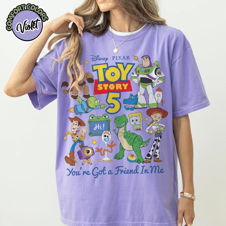 May include: A light purple short-sleeve t-shirt featuring a graphic print of Disney Pixar Toy Story characters including Woody, Buzz Lightyear, Jessie, Rex, and Forky. The text reads Toy Story 5 and You've Got a Friend In Me with a small logo.