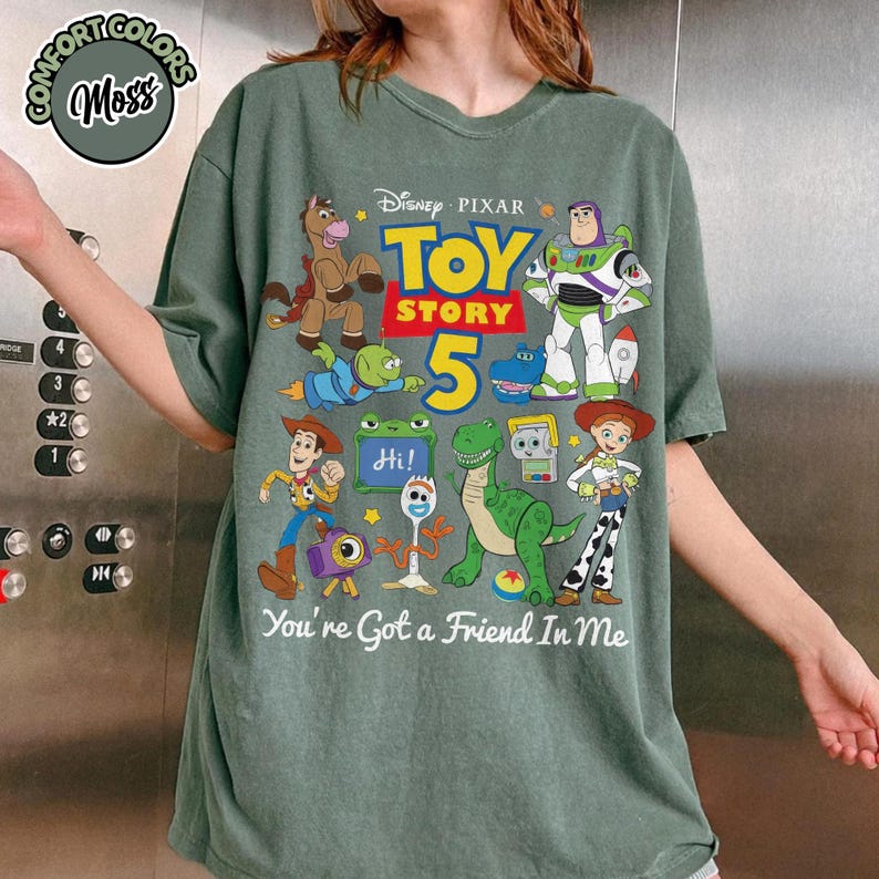 May include: A moss green t-shirt featuring a graphic print of Disney Pixar Toy Story 5 characters including Woody, Buzz Lightyear, Jessie, Rex, and Forky. The text reads Toy Story 5 and You've Got a Friend In Me. A Comfort Colors Moss logo is in the corner.