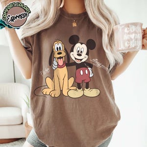 May include: Brown t-shirt featuring a graphic of Mickey Mouse and Pluto. Mickey Mouse is wearing red shorts and yellow shoes. Pluto is a light yellow color with a red collar. The shirt has the text "Mickey Mouse" and "Pluto".