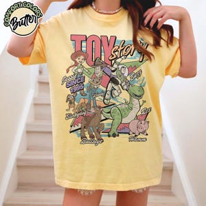 May include: Yellow t-shirt featuring a graphic of Toy Story characters, including Woody, Buzz Lightyear, and Jessie. The design includes the text "Toy Story" and "Since 1995". The shirt has short sleeves and a crew neck.