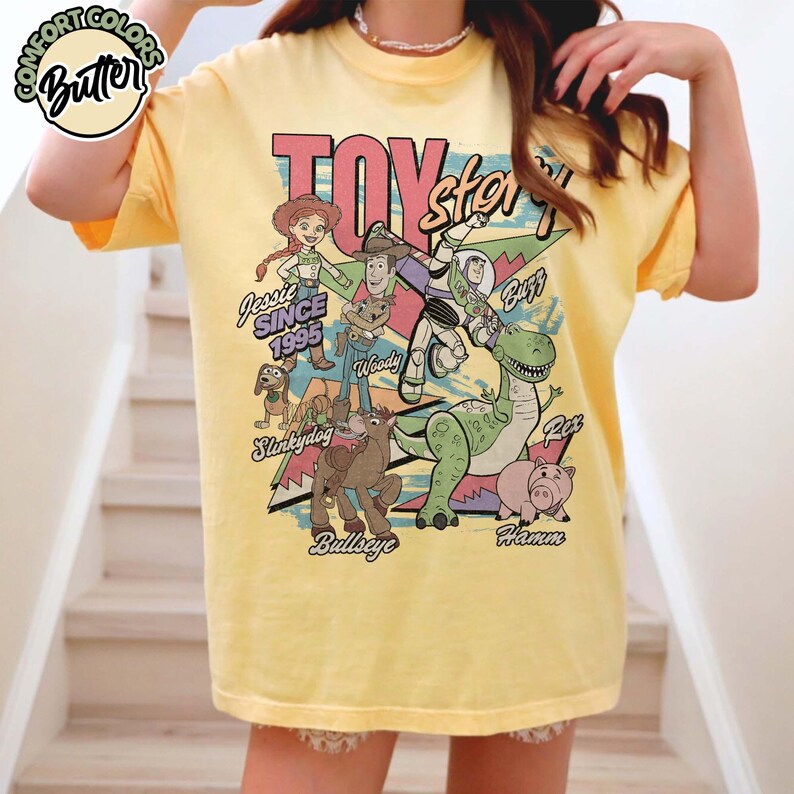 May include: Yellow t-shirt featuring a graphic of Toy Story characters, including Woody, Buzz Lightyear, and Jessie. The design includes the text "Toy Story" and "Since 1995". The shirt has short sleeves and a crew neck.