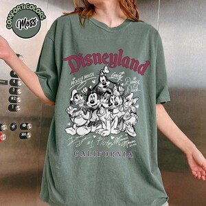 Puede incluir: Camiseta verde Comfort Colors con un gr&aacute;fico de Disneyland con Mickey Mouse, Goofy, Donald Duck, Daisy Duck, Pluto y Minnie Mouse. El texto "Disneyland California" est&aacute; impreso en color burdeos.