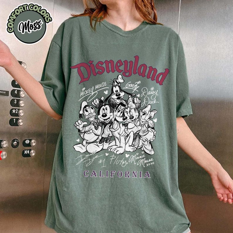 Puede incluir: Camiseta verde Comfort Colors con un gr&aacute;fico de Disneyland con Mickey Mouse, Goofy, Donald Duck, Daisy Duck, Pluto y Minnie Mouse. El texto "Disneyland California" est&aacute; impreso en color burdeos.