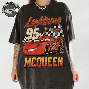 Pu&ograve; includere: T-shirt grigio scuro con una grafica di Saetta McQueen del film Cars. Il design include il testo "Lightning 95 McQueen" in rosso e giallo, con una bandiera a scacchi e un trofeo.