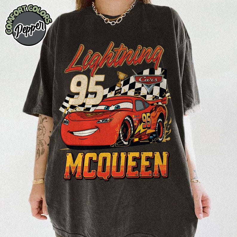 Pu&ograve; includere: T-shirt grigio scuro con una grafica di Saetta McQueen del film Cars. Il design include il testo "Lightning 95 McQueen" in rosso e giallo, con una bandiera a scacchi e un trofeo.