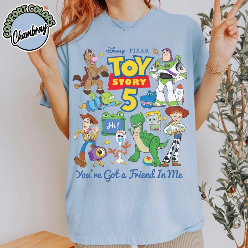 May include: A light blue t-shirt featuring a colorful graphic of Disney Pixar Toy Story characters including Woody, Buzz Lightyear, Jessie, Rex, and Forky. The design includes the text Toy Story 5 and You're Got a Friend In Me with a small logo.