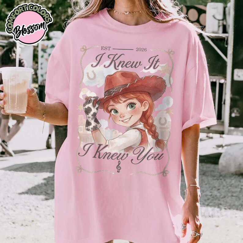 Peut inclure: Un t-shirt rose clair avec une illustration de personnage portant un chapeau de cow-boy marron et un gant &agrave; motif vache. Le texte indique "EST 2026", "I Knew It" et "I Knew You" avec une note de musique. Le motif inclut des nuages et des fers &agrave; cheval.