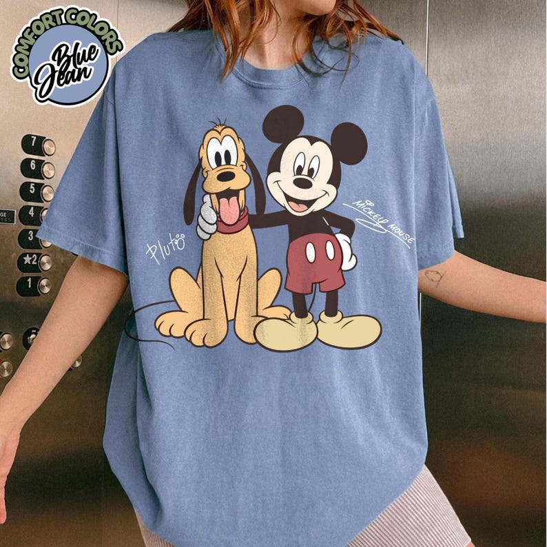May include: Blue jean comfort colors t-shirt featuring Mickey Mouse and Pluto. Mickey Mouse is wearing red shorts and yellow shoes. Pluto is a tan color with a red tongue. The shirt has the words "Mickey Mouse" and "Pluto" printed on it.