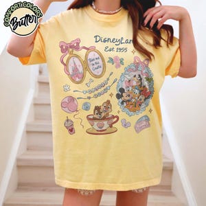 Peut inclure: T-shirt jaune avec des graphismes de Disneyland. Le t-shirt pr&eacute;sente un n&oelig;ud rose, un m&eacute;daillon avec la Tour Eiffel et une tasse &agrave; th&eacute; avec Tic et Tac. Le texte "Disneyland Est. 1955" est &eacute;galement visible.