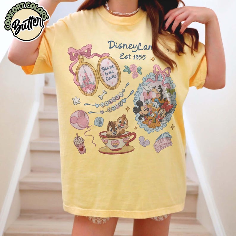 Peut inclure: T-shirt jaune avec des graphismes de Disneyland. Le t-shirt pr&eacute;sente un n&oelig;ud rose, un m&eacute;daillon avec la Tour Eiffel et une tasse &agrave; th&eacute; avec Tic et Tac. Le texte "Disneyland Est. 1955" est &eacute;galement visible.
