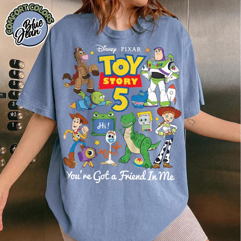 May include: A blue jean colored t-shirt featuring a graphic print of Disney Pixar Toy Story 5 characters including Woody, Buzz Lightyear, Jessie, Rex, and Forky. The text reads Toy Story 5 and You've Got a Friend In Me. A Comfort Colors logo is visible.