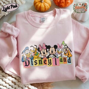 May include: A light pink crewneck sweatshirt featuring an embroidered graphic of Disney characters including Donald Duck, Pluto, Minnie Mouse, Mickey Mouse, Daisy Duck, and Goofy. Below the characters, the text Disneyland is embroidered in black.