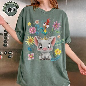 May include: Green t-shirt featuring a cartoon pig and rooster with colorful flowers. The shirt has the words "Comfort Colors Moss" in a circular design at the top left.