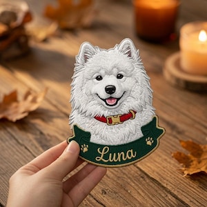 May include: A white Samoyed dog embroidered patch featuring a red collar with a gold buckle. Below the dog is a dark green banner with gold embroidered paw prints and the name Luna written in a cursive gold script font on the patch.