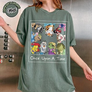 Puede incluir: Camiseta verde salvia con un gr&aacute;fico rectangular con personajes de dibujos animados de varias pel&iacute;culas. El texto "Once Upon A Time" est&aacute; impreso debajo. La camiseta es de la marca Comfort Colors.