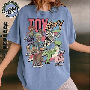 May include: Blue jean-colored t-shirt featuring a graphic of Toy Story characters, including Woody, Buzz, and Rex. The design includes the text "TOY story" and "Jessie Since 1995". The shirt has a round neckline and short sleeves.