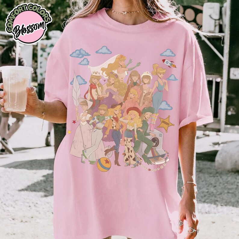 May include: A light pink t-shirt featuring a large graphic print of multiple animated female characters in various outfits, including a cowgirl, a space ranger, and a princess, surrounded by clouds, a rocket, and a star, with a circular logo in the corner.