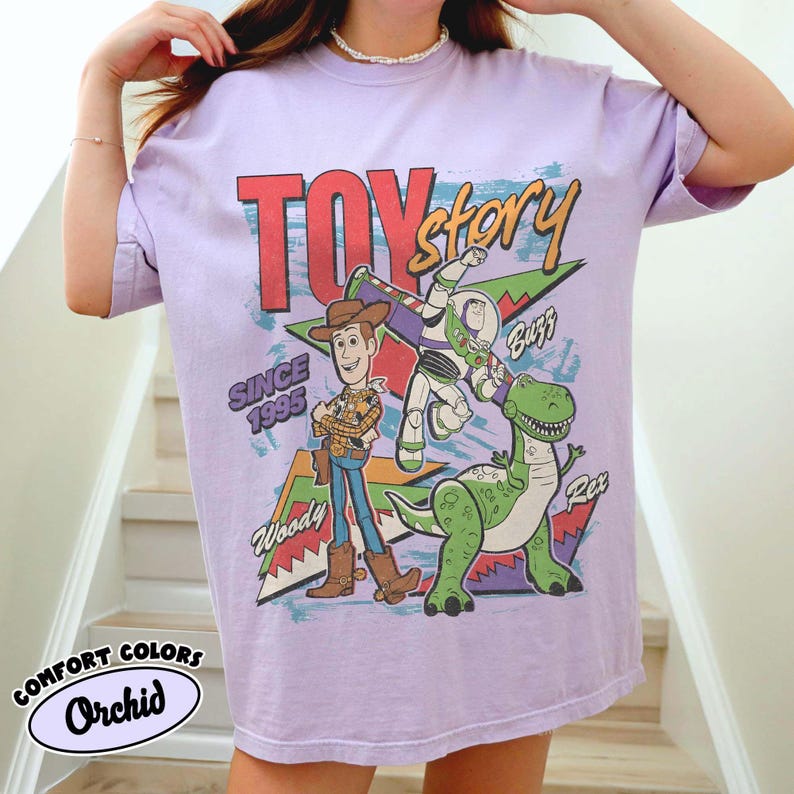 Puede incluir: Camiseta lila con la frase "TOY Story" en rojo y blanco, con Woody, Buzz Lightyear y Rex. La camiseta tambi&eacute;n incluye el texto "SINCE 1995" y "Comfort Colors Orchid".