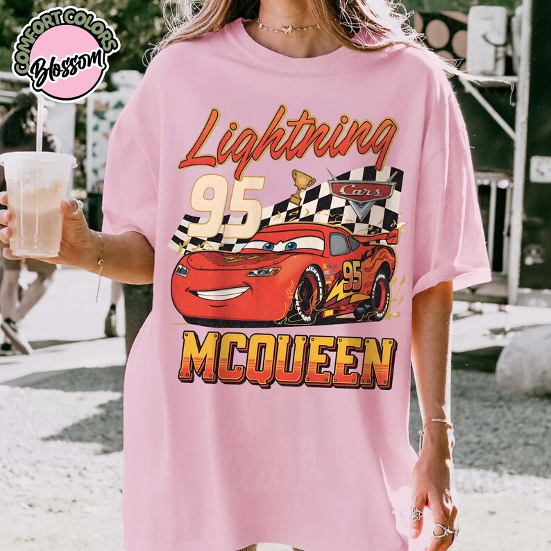 Pu&ograve; includere: T-shirt rosa chiaro con una grafica di Saetta McQueen del film Cars. Il design include il testo "Lightning 95" e "McQueen" in stile vintage. Sono inclusi anche una bandiera a scacchi e un trofeo.