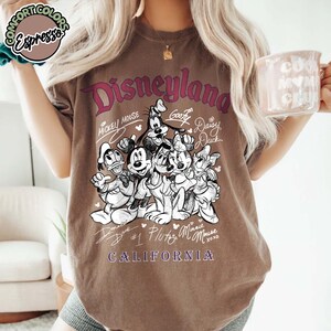 Puede incluir: Camiseta marr&oacute;n con un gr&aacute;fico de personajes de Disney, incluyendo Mickey Mouse y Goofy. La palabra "Disneyland" est&aacute; impresa encima de los personajes, con "California" debajo. La camiseta tiene un aspecto vintage.
