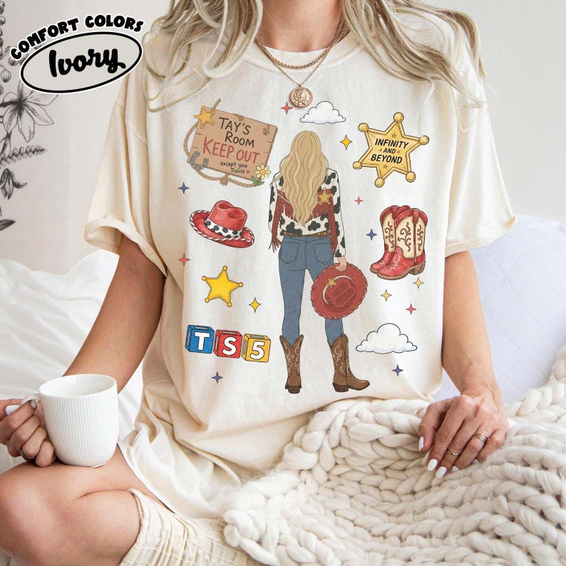 May include: An ivory short-sleeve t-shirt featuring a graphic print of a person with long blonde hair, wearing a cow-print jacket, blue jeans, and brown boots. The design includes a wooden sign reading Tay's Room Keep Out, a sheriff star, red cowboy hats, and blocks labelled TS5.