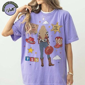 May include: A light purple short-sleeve t-shirt featuring a graphic of a person with long blonde hair wearing a cow-print jacket, blue jeans, and brown boots. The design includes a red cowboy hat, a sheriff star labelled Infinity and Beyond, a wooden sign reading Tay's Room Keep Out, and blocks spelling TS 5.