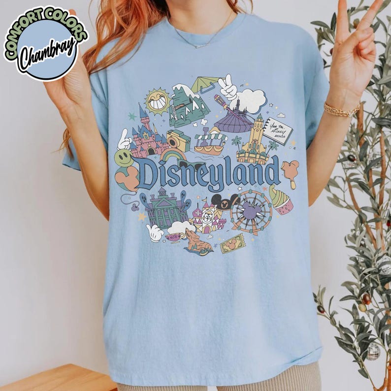 Puede incluir: Camiseta azul claro con un gr&aacute;fico de Disneyland. El dise&ntilde;o incluye ilustraciones de un castillo, una monta&ntilde;a rusa y otros iconos del parque. El texto "Disneyland" se muestra en una fuente caprichosa. El logotipo de Comfort Colors y Chambray est&aacute; en la esquina superior izquierda.