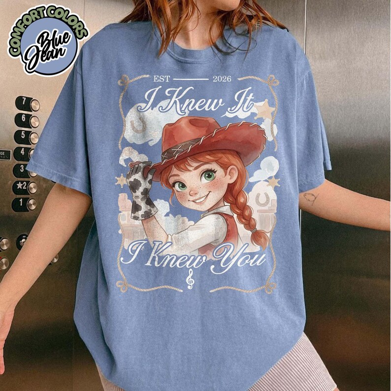 Peut inclure: Un t-shirt bleu &agrave; manches courtes avec l'illustration d'une cow-girl de dessin anim&eacute; aux cheveux roux, portant un chapeau marron et un gant &agrave; motifs de vache. Le texte indique "EST 2026", "I Knew It" et "I Knew You" avec une note de musique.