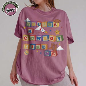 May include: A berry-colored t-shirt featuring a graphic of colorful alphabet blocks spelling out the text "YOU'RE A COWBOY LIKE ME". The design includes illustrations of a rocket, a sheriff star, a cowboy hat, cowboy boots, and a ball.