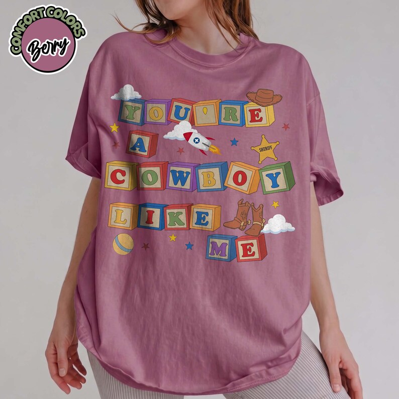 May include: A berry-colored t-shirt featuring a graphic of colorful alphabet blocks spelling out the text "YOU'RE A COWBOY LIKE ME". The design includes illustrations of a rocket, a sheriff star, a cowboy hat, cowboy boots, and a ball.