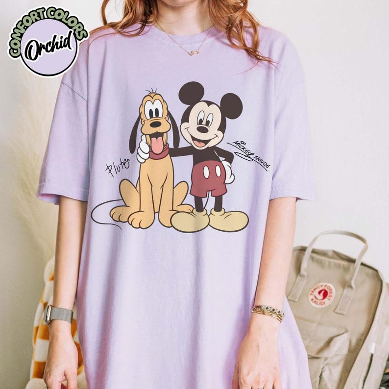 May include: Orchid Comfort Colors lilac t-shirt featuring a graphic of Mickey Mouse and Pluto. Mickey Mouse is wearing red shorts and Pluto is a tan color. The text "Pluto" and "Mickey Mouse" are also visible.